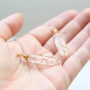 May include: Two clear quartz crystal pendants, each wrapped in gold-coloured wire. The crystals are elongated and pointed, with a loop at the top for hanging. The wire is intricately woven around the crystals, creating a cage-like design. The pendants are displayed in an open hand.