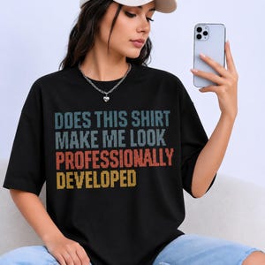 May include: A black short-sleeve t-shirt featuring pixelated text in blue, orange, and yellow. The graphic reads: DOES THIS SHIRT MAKE ME LOOK PROFESSIONALLY DEVELOPED. The shirt has a crew neckline and a loose, relaxed fit with short sleeves.