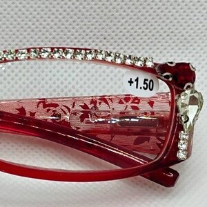 May include: Red reading glasses with a +1.50 magnification. The frames are adorned with rhinestones along the top and at the temples. The lenses are clear, and the arms have a floral pattern.