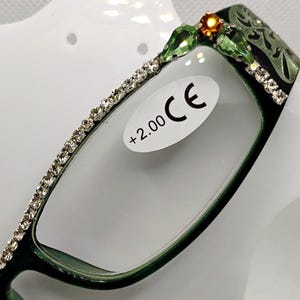 May include: Green-framed reading glasses with rhinestone accents along the top and sides. The upper frame features a decorative design with green and orange gemstones. The lenses have a +2.00 magnification and a CE marking.