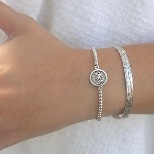 May include: A silver beaded bracelet featuring a circular charm with a relief of a figure, paired with a silver cuff bracelet engraved with a floral pattern. Both pieces are metallic silver in color and worn together on the wrist.