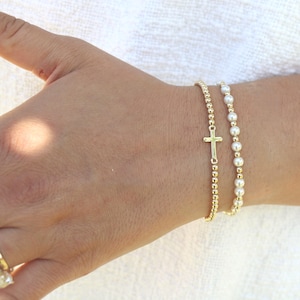 May include: A set of two bracelets worn on the wrist. One bracelet features small gold-toned beads with a central gold-toned cross charm. The second bracelet consists of a single strand of white spherical beads interspersed with small gold-toned beads.