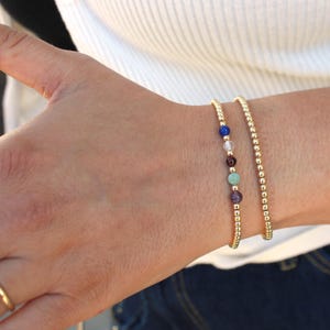 May include: Two gold beaded bracelets. One bracelet consists entirely of small, round, metallic gold beads. The second bracelet features a series of small, round, metallic gold beads interspersed with five distinct, smooth, multi-colored gemstone beads.