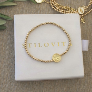 May include: A gold beaded bracelet with a circular charm featuring an embossed design, resting on a white box. The box has the word "STILOVITA" printed in gold. Additional gold jewelry is visible in the background.