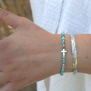May include: A beaded bracelet featuring round turquoise-colored stones and small silver-toned metallic beads with a central silver-toned cross charm, paired with a wide silver-toned cuff bracelet featuring an engraved floral scroll pattern.