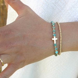 Può includere: Due braccialetti su un polso. Uno presenta perline turchesi e un ciondolo a forma di croce dorata. L'altro è un delicato braccialetto di perline dorate. I braccialetti sono indossati su una carnagione chiara.