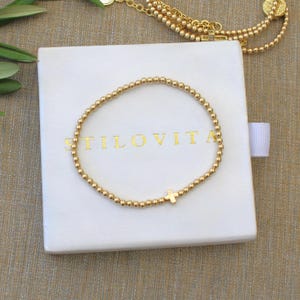 May include: Gold beaded bracelet with a small cross charm, resting on a white box with the word "STILOVITA" printed in gold. A gold necklace is visible in the background. The bracelet is a delicate piece of jewelry.