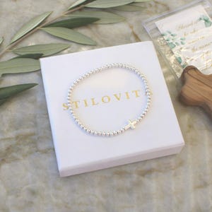 May include: A silver beaded bracelet featuring a small silver cross charm, resting on a white square box with the gold text STILOVITA printed on the centre. The bracelet is composed of small, uniform metallic spheres arranged in a circular loop.