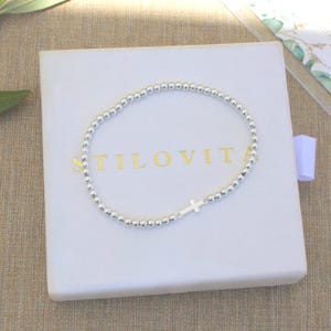 Puede incluir: Pulsera de cuentas plateadas con un pequeño dije de cruz, presentada en una caja blanca con letras doradas "STILOVITA". La pulsera está hecha de pequeñas cuentas redondas plateadas.