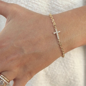 May include: A gold-colored bracelet with a cross pendant. The cross is encrusted with small, clear gemstones. The bracelet is on a wrist, and the chain links are visible. The bracelet is a delicate piece of jewelry.