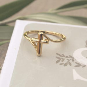 May include: A polished gold-tone ring featuring a central cross design integrated into the band. The metal has a smooth, reflective surface with a continuous, flowing shape that forms the cross symbol as the primary focal point of the jewellery piece.