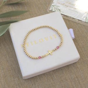 May include: Gold beaded bracelet with a cross charm and pink accents, presented on a white box. The box has the word "STILOVIT" printed on it. A small card with text is visible in the background.