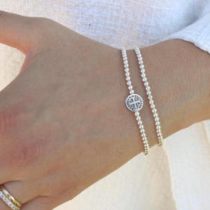 Pode incluir: Duas pulseiras de contas prateadas no pulso. Uma pulseira apresenta um pequeno pingente circular com um design de cruz em relevo e texto, enquanto a outra é um fio simples de contas metálicas uniformes. Um anel dourado também é visível.