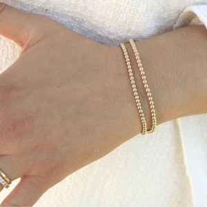 May include: A gold beaded bracelet is worn on a wrist. The bracelet is made of small, round beads strung together. The bracelet is a delicate piece of jewelry, perfect for everyday wear. The bracelet is a simple yet elegant accessory.