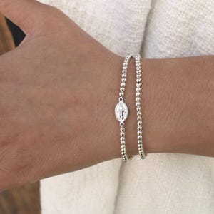 Puede incluir: Dos pulseras de cuentas plateadas. Una pulsera presenta un pequeño colgante ovalado con una figura religiosa. Las pulseras se llevan en la muñeca y las cuentas son pequeñas y redondas.