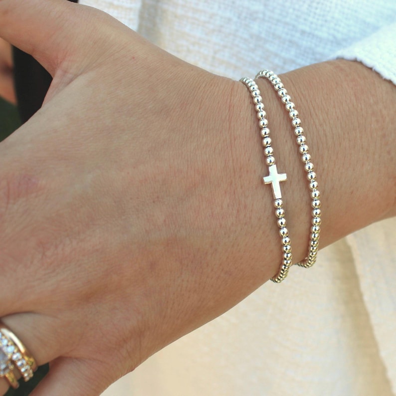 May include: Two silver beaded bracelets are worn on a wrist. One bracelet features a small cross charm. The bracelets are made of small, round beads. The background is a light color.