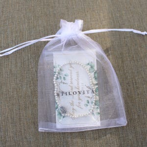 May include: A pearl bead bracelet with a silver charm is displayed inside a white organza gift bag. The bag contains a card with text and a floral design. The text on the card reads "May the Lord reward you for your kindness."