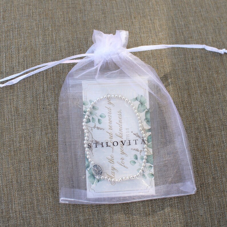 May include: A pearl bead bracelet with a silver charm is displayed inside a white organza gift bag. The bag contains a card with text and a floral design. The text on the card reads "May the Lord reward you for your kindness."