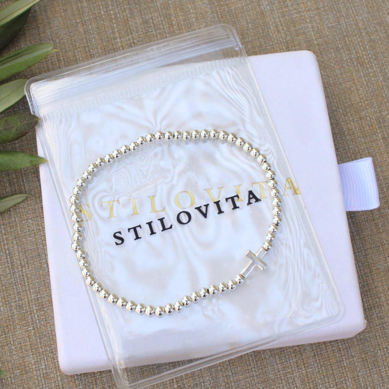 May include: A silver beaded bracelet with a cross charm, displayed in a clear plastic bag with the brand name "STILOVITA" printed on it. The bracelet rests on a white box with a ribbon.