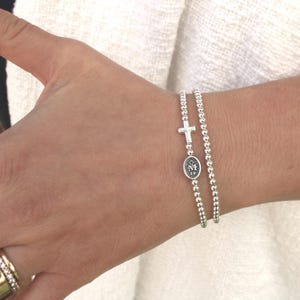 May include: Two silver beaded bracelets. One bracelet features a small cross charm, and the other has an oval charm with an emblem. The bracelets are worn on a wrist.