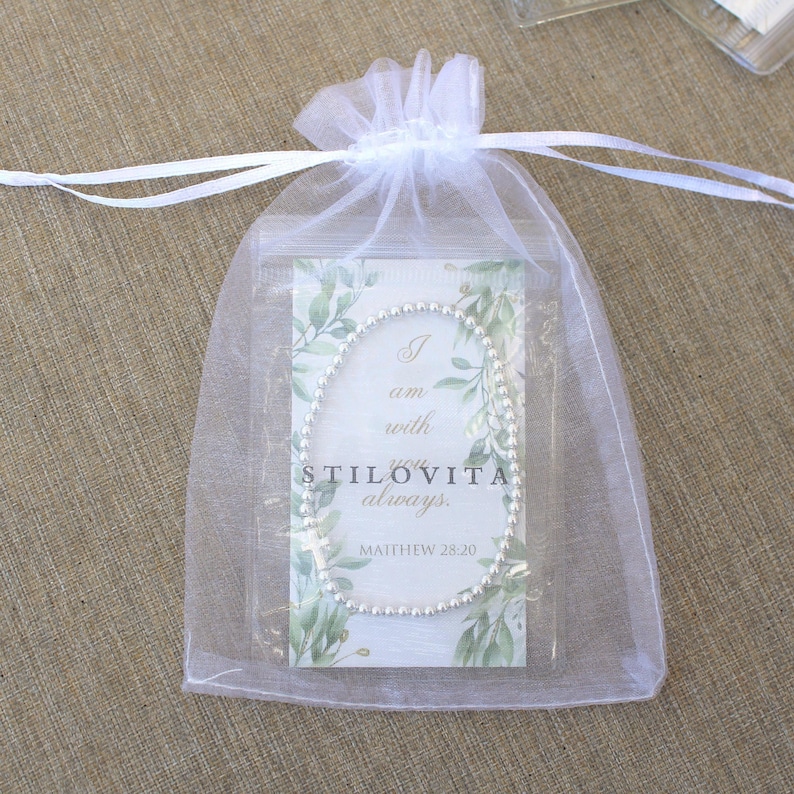 May include: A silver beaded bracelet inside a clear plastic bag, enclosed in a white organza drawstring bag. The card inside the bag reads "I am with you always. STILOVITA. Matthew 28:20" with a green leaf design.