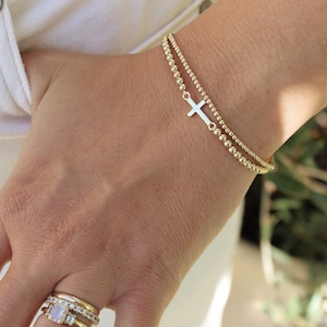 May include: A gold bracelet with two strands of small beads. One strand features a small cross charm. The bracelet is worn on a wrist, and the image also shows several gold rings on a finger.