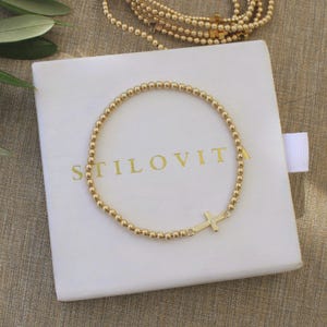May include: A gold beaded bracelet with a cross charm rests on a white box. The box has the word "STILOVITA" printed in gold. Additional gold beaded jewelry is visible in the background.