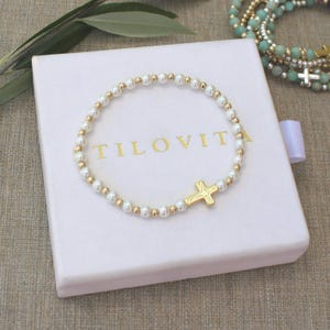 May include: A beaded bracelet featuring alternating white pearls and small gold-toned spheres, centered with a gold-toned cross charm. The bracelet rests on a white square gift box with the gold-printed text TILOVITA centered on the lid.