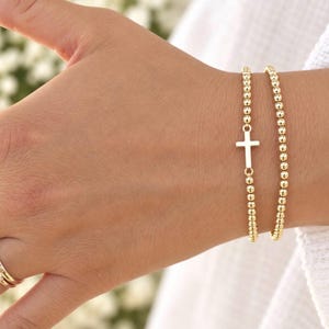 May include: A set of two gold-toned beaded bracelets worn on the wrist. One bracelet features a small, centered gold-toned cross charm. The beads are uniform in size and round, creating a simple, continuous chain design on each bracelet.