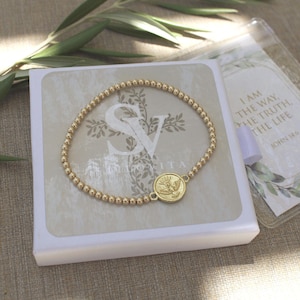 May include: A gold-toned beaded bracelet featuring a circular charm with a dove emblem. The bracelet is displayed on a white square box with the text SV and I AM THE WAY, THE TRUTH, THE LIFE JOHN 14:6 printed on the packaging surface.