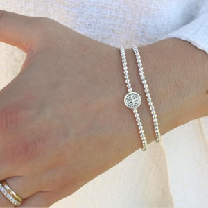 May include: Two silver beaded bracelets, each made of small, round beads, with a round charm featuring a cross design. The bracelets are worn on a wrist. A gold ring is visible on a finger.