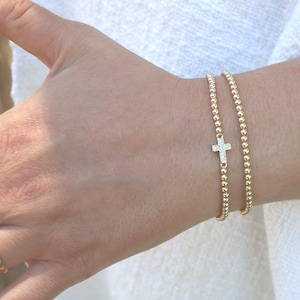 May include: A gold-toned beaded bracelet featuring a small, central cross charm encrusted with clear, sparkling stones. The bracelet consists of uniform, polished spherical beads strung together, creating a delicate and reflective metallic appearance.