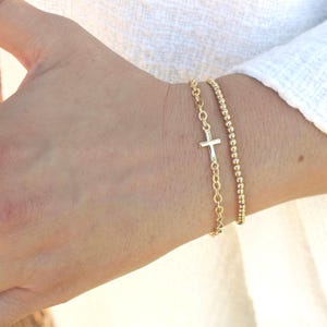 May include: Two gold bracelets. One features a cross pendant on a chain, the other is a beaded design. The bracelets are worn on a wrist, against a white background.