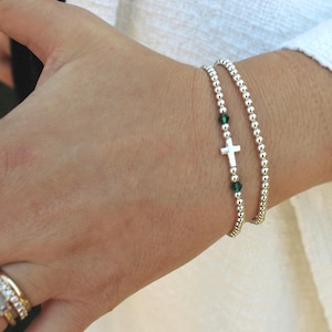 Puede incluir: Dos pulseras de cuentas plateadas. Una pulsera presenta una pequeña cruz plateada y cuentas verdes. La otra pulsera está hecha de pequeñas cuentas plateadas. Las pulseras se llevan en la muñeca.
