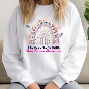 May include: White sweatshirt with a rainbow design and the text "I love someone rare. Rare Disease Awareness".