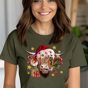 May include: A green t-shirt with a cartoon image of a Highland cow wearing a Santa hat and Christmas lights. The cow is holding a gift and has a red nose.