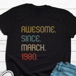 May include: Black t-shirt with a vintage-style graphic that reads "Awesome. Since. March. 1980."