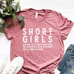 May include: A pink t-shirt with the text "SHORT GIRLS" in white. The text below reads "GOD ONLY LETS THINGS GROW UNTIL THEY'RE PERFECT. SOME OF US DIDN'T TAKE AS LONG AS OTHERS."