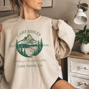 May include: A beige sweatshirt with a green graphic design of a mountain range, lake, and trees. The text "Camp Ashley Est. 2024 Ashley's Bachelorette Lake Tahoe, CA" is printed on the sweatshirt.