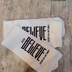 May include: Two white socks with the words "I Believe God" and "Believe God" printed in black.