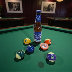 May include: A bottle of beer with a blue and white label, and several billiard balls on a green pool table. The bottle is labelled "White Tide". The balls are numbered 2, 12, 13, and 16.