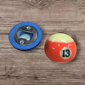 May include: A bottle opener with a blue circular base and a metal insert, positioned next to a billiard ball numbered 13. The billiard ball is cream-colored with a wide orange band around the center and the black number 13 printed on a white circle.