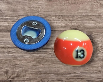 Puede incluir: Un abridor de botellas azul con base circular e inserto metálico, junto a una bola de billar redonda con parte superior color crema, banda central naranja y el número 13 negro impreso sobre un fondo circular blanco.