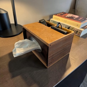 May include: A wooden tissue box with a two-tone design. The box is rectangular with a dark brown base and a lighter brown top. A remote control and a black device sit in a recessed area on top. White tissues are pulled from a circular opening.