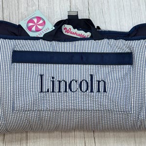 May include: A navy blue and white striped personalized picnic blanket with the name "Lincoln" embroidered on the front. Features a navy blue pocket and trim, with a handle and a "Washable!" tag.