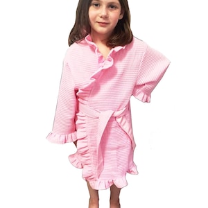 May include: A pink waffle-textured robe with ruffled edges and a matching belt. The robe has a wrap-around design and is worn by a person. The robe's color is a soft, light pink.