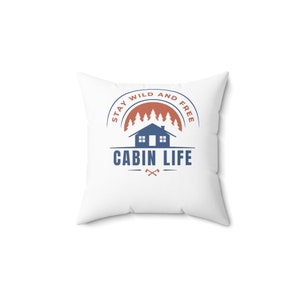 May include: White throw pillow with a blue and red logo that says "Stay Wild and Free Cabin Life". The logo features a cabin with a red sunset behind it and pine trees.