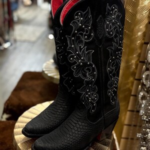 May include: Black cowboy boots featuring a textured snakeskin-patterned foot and a tall shaft with intricate black embroidery and silver stud accents. The interior lining of the boots is a vibrant red, and they have a pointed toe and a stacked heel.