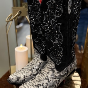 May include: A pair of tall western-style boots featuring a black suede upper shaft with white embroidered floral scrollwork and silver stud accents. The foot section is textured with a grey and black snakeskin pattern, and the interior is red.