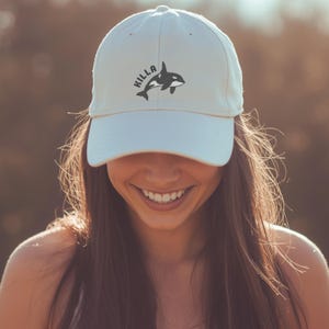 May include: A pale baseball cap featuring the word "Hilla" and a black and white orca whale design. The cap is worn by a person with long brown hair, smiling. The background is blurred with warm colours.